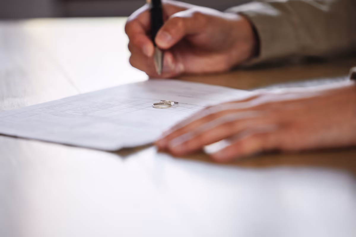 Hands signing divorce papers with wedding rings on the page